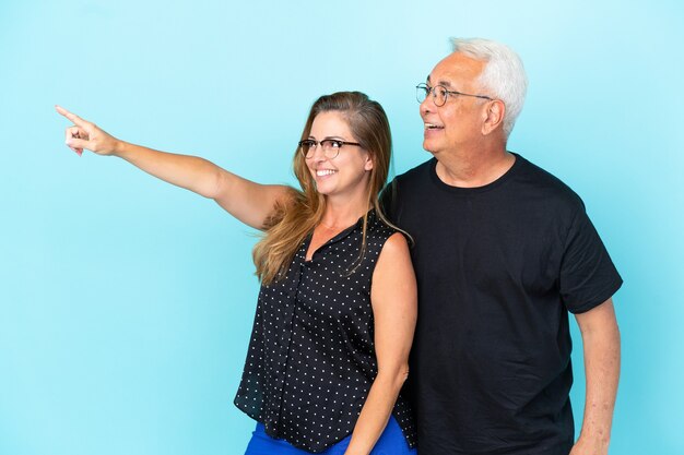 Casal de meia-idade isolado em fundo azul apontando o dedo para o lado e apresentando um produto