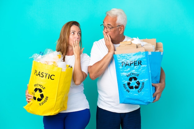 Casal de meia-idade segurando um saco de reciclagem cheio de papel e plástico isolado no fundo branco, cobrindo a boca com as mãos por dizer algo impróprio