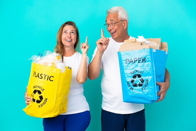 Casal de meia-idade segurando um saco de reciclagem cheio de papel e plástico isolado no fundo branco com a intenção de descobrir a solução enquanto levanta um dedo