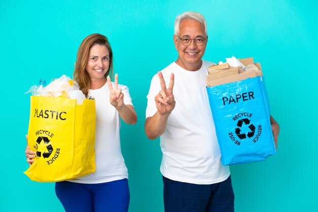 Casal de meia-idade segurando um saco de reciclagem cheio de papel e plástico isolado no fundo branco, sorrindo e mostrando o sinal da vitória