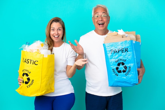 Casal de meia-idade segurando uma sacola de reciclagem cheia de papel e plástico isolado no fundo branco com expressão facial surpresa e chocada