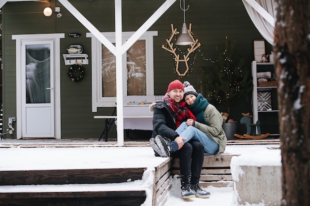 Casal de meia idade sentado em um terraço nevado passo em frente da casa, posando para uma foto. Ambos usando chapéus, jaquetas e lenços. Ele está segurando os tornozelos dela sobre os joelhos.