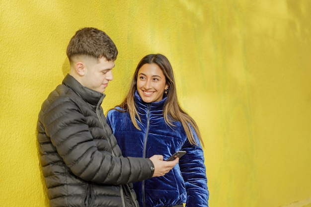 Casal de menino e menina encostado em uma parede amarela assistindo o celular com espaço de cópia