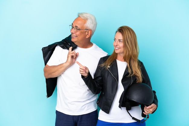 Casal de motociclistas de meia-idade com um capacete de motociclista isolado em um fundo azul apresentando uma ideia enquanto olha sorrindo para