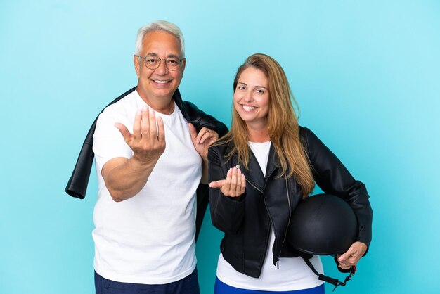 Casal de motociclistas de meia-idade com um capacete de motociclista isolado em um fundo azul, convidando a vir com a mão. feliz que você veio