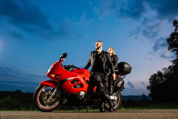 Casal de motoqueiros legal em jaquetas de couro sentado na bicicleta esportiva vermelha ao ar livre