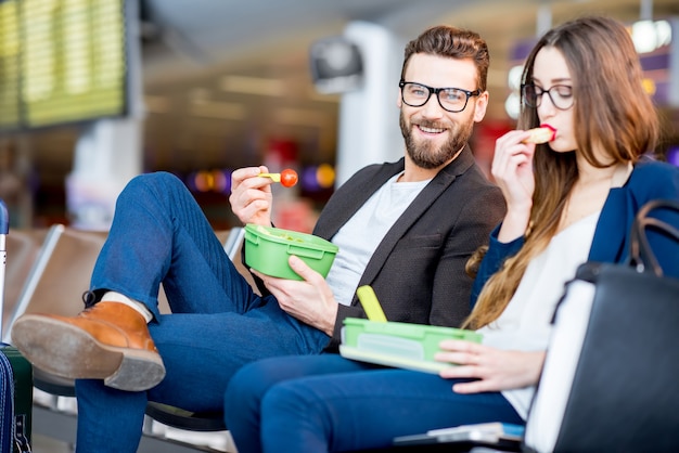 Casal de negócios elegantes comendo com lancheiras, sentado na sala de espera no aeroporto. fazendo um lanche durante a viagem de negócios