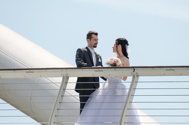 Casal de noiva de pé junto à grade da ponte contra o céu claro