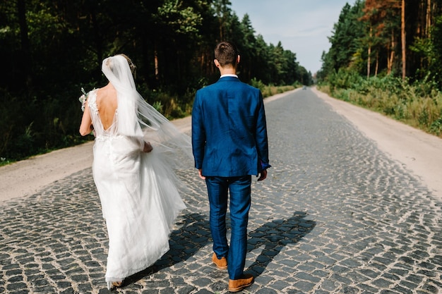 Casal de noivos andando na natureza A noiva com buquê e noivo vão na estrada pavimentada e um passeio pela floresta Recém-casados ao ar livre Vista traseira