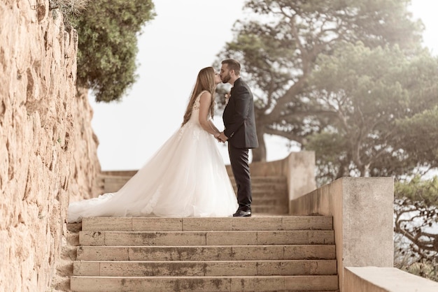 Casal de noivos beijando e abraçando nas escadas com árvores verdes ao fundo