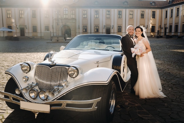 Casal de noivos elegantes no pátio do castelo perto de um carro retrô