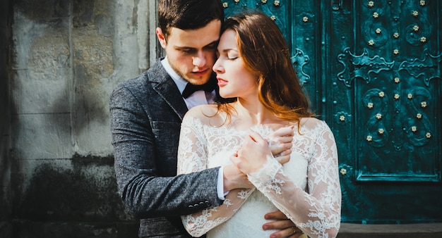 Casal de noivos se abraça perto da porta verde vintage. Paredes de pedra no fundo da cidade antiga. Noiva rústica com cabelo solto em vestido de renda e noivo de terno cinza e gravata borboleta. Abraço terno. Amor romântico.