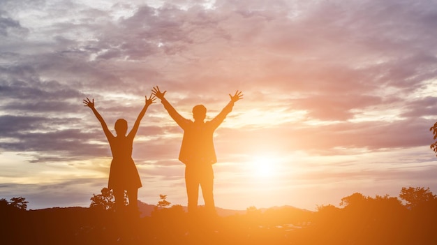 Casal de silhueta com os braços levantados de pé contra o céu nublado durante o pôr do sol