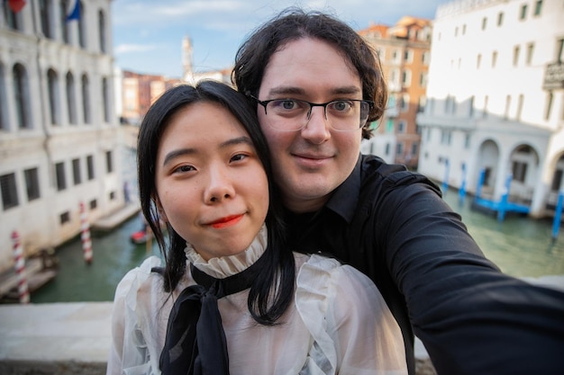 Casal de turistas multirraciais visitando Veneza tira uma selfie na ponte e no canal ao fundo