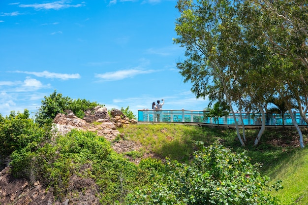 Casal de turistas tirando fotos em um mirante na montanha. Há um lindo mar azul ao fundo.