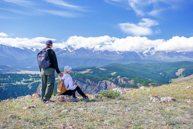 Casal de viajantes homem e mulher sentada na falésia relaxante montanhas. caminhantes com mochilas relaxando no topo de uma montanha e apreciando a vista do vale.