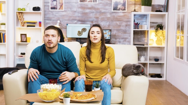 Casal decepcionado depois que seu time de futebol favorito perdeu o jogo. Gato deitado no sofá. Pipoca, pizza e refrigerante na mesa de centro.