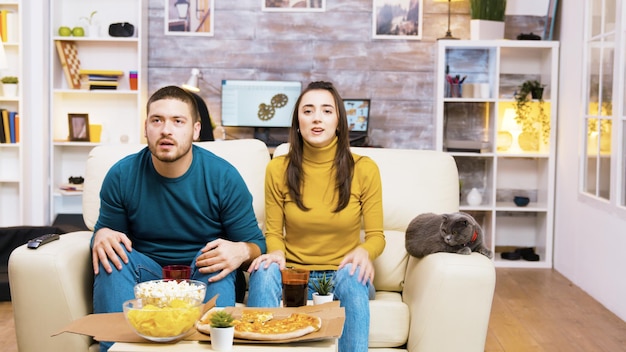 Casal decepcionado depois que seu time de futebol favorito perdeu o jogo. gato deitado no sofá. pipoca, pizza e refrigerante na mesa de centro.