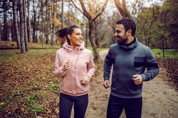 Casal dedicado positivo sorridente no sportswear, olhando um para o outro e correndo na natureza