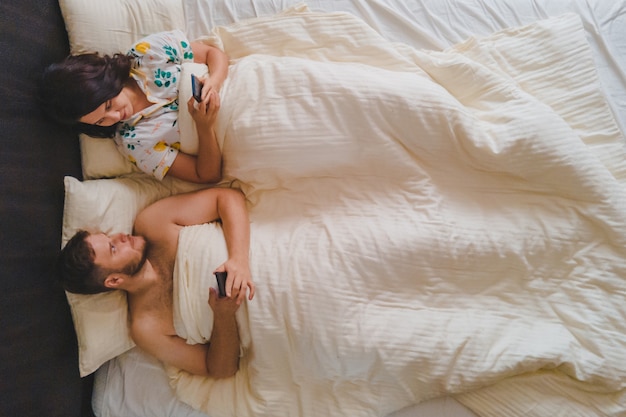 Casal deitado na cama, surfando na internet no vício moderno de telefones. ver diretamente acima