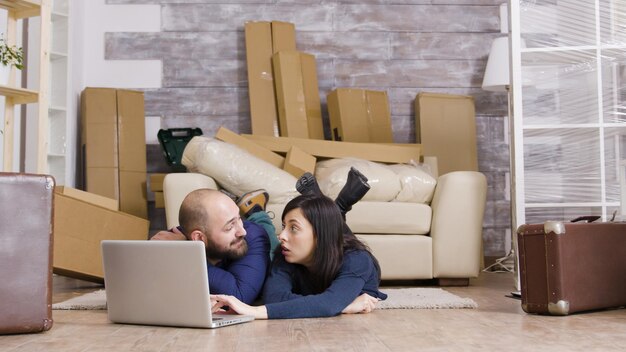 Casal deitado no chão de seu novo apartamento, fazendo compras online no laptop. amplie a imagem.