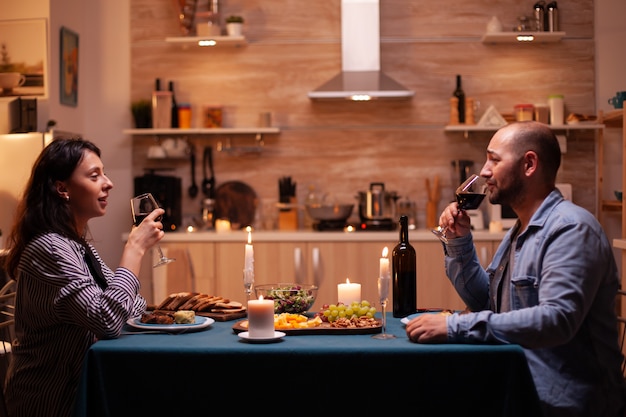 Casal desfrutando de uma taça de vinho. relaxe pessoas felizes tilintando, sentadas à mesa na cozinha, apreciando a refeição, comemorando aniversário na sala de jantar.