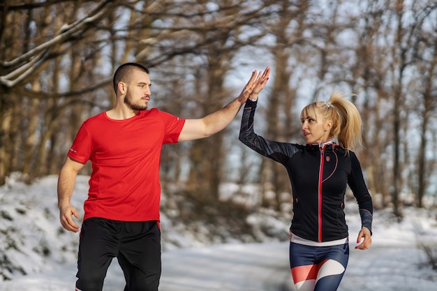 Casal desportivo correndo juntos na neve na natureza no inverno e dando high five um ao outro wi ...