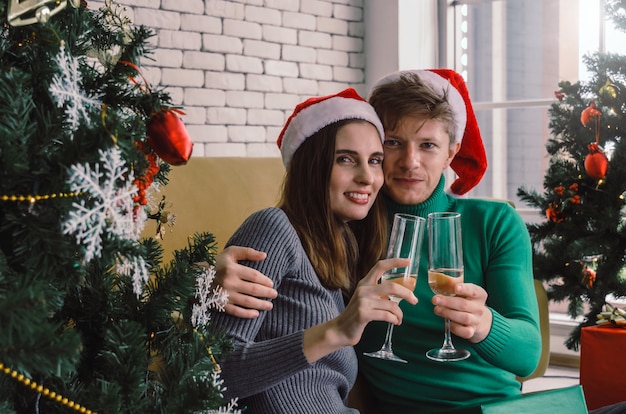 Casal doce caucasiano com chapéu de Papai Noel vermelho gosta de beber champanhe e olhando com árvore de Natal comemorando em casa