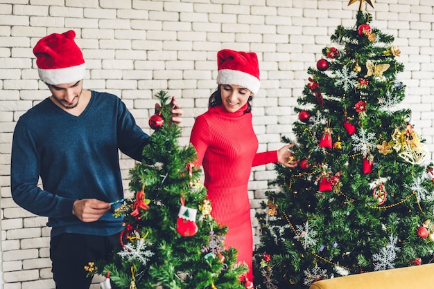 Casal doce romântico em gorros se divertindo decorando a árvore de Natal e sorrindo enquanto comemorava a véspera de ano novo