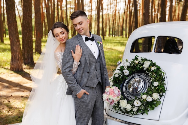 Casal elegante casamento, noiva e noivo abraçando perto de carro recém casado branco com decoração de coroa de flores com flores frescas