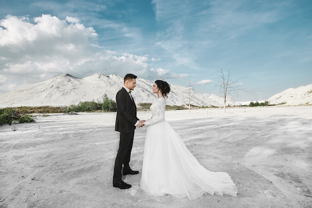 Casal elegante e bonito, garota modelo morena sexy e bonita com penteado elegante, vestido de renda branca e homens bonitos elegantes terno moderno posando ao ar livre no deserto de sais