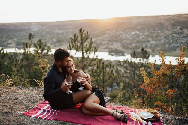 Casal elegante em roupas pretas fazendo um piquenique ao ar livre com vinho ao pôr do sol