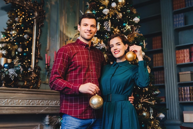 Foto casal em trajes festivos segurando ornamentos de natal dourados de pé junto a uma árvore ricamente decorada em uma sala elegante