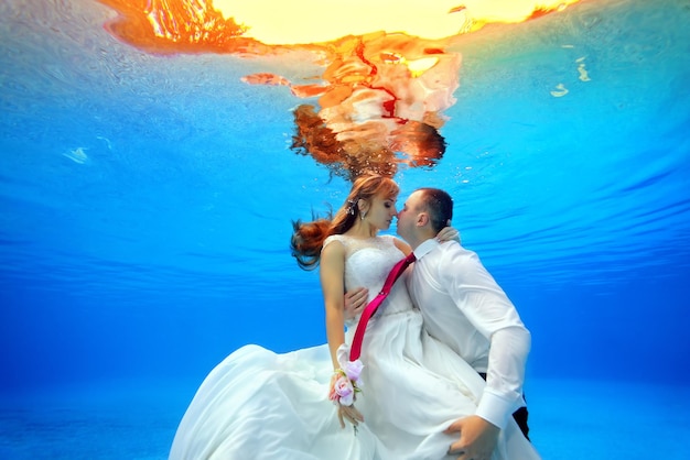 Foto casal em vestidos de noiva abraçando e beijando debaixo d'água na piscina ao pôr do sol