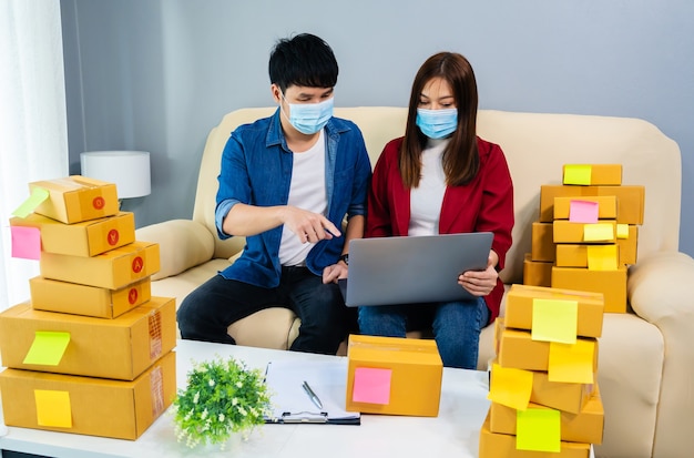 Casal empresário trabalhando com um laptop para preparar a caixa de pacotes para entrega ao cliente no escritório doméstico, pessoas usando uma máscara facial para proteger a pandemia de coronavírus