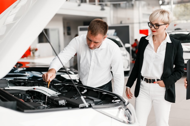 Casal escolhe um carro na concessionária. Homem que olha sob o capô do carro.