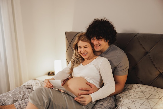 Casal esperando feliz olhando para exame médico de seu bebê enquanto relaxa em uma cama no quarto.