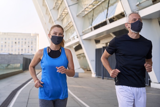 Casal esportivo de meia-idade, homem e mulher, usando máscaras protetoras e correndo juntos em meio urbano