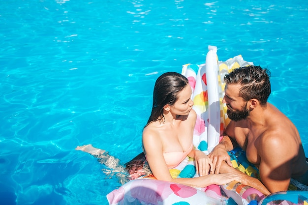 Casal está de pé na piscina e nos olha. Eles estão molhados. Casal se inclina para colchão de ar.