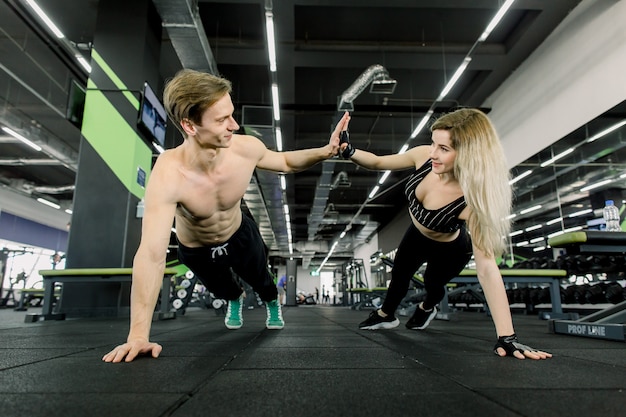 Casal fazendo flexões no treinamento no ginásio. Jovem casal desportivo malhando juntos em uma academia. Fazendo exercícios de prancha enquanto se abraçam por uma mão.