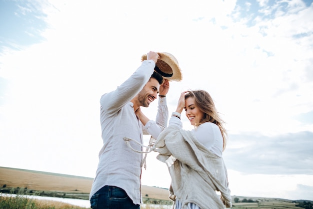 Foto casal feliz apaixonado, abraçando, beijando e sorrindo contra o céu em campo. chapéu na mão de menina