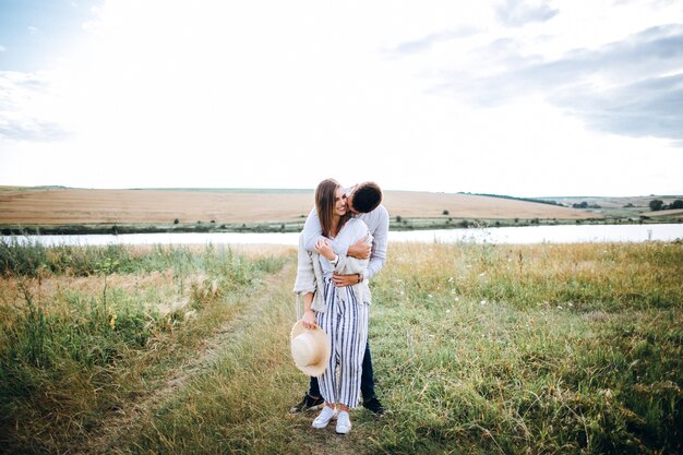 Foto casal feliz apaixonado, abraçando, beijando e sorrindo contra o céu em campo. chapéu na mão de menina