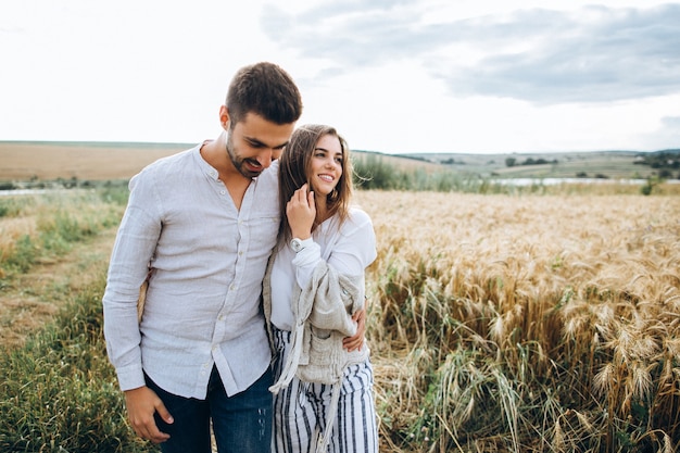 Casal feliz apaixonado, abraçando, beijando e sorrindo contra o céu em campo. Chapéu na mão de menina