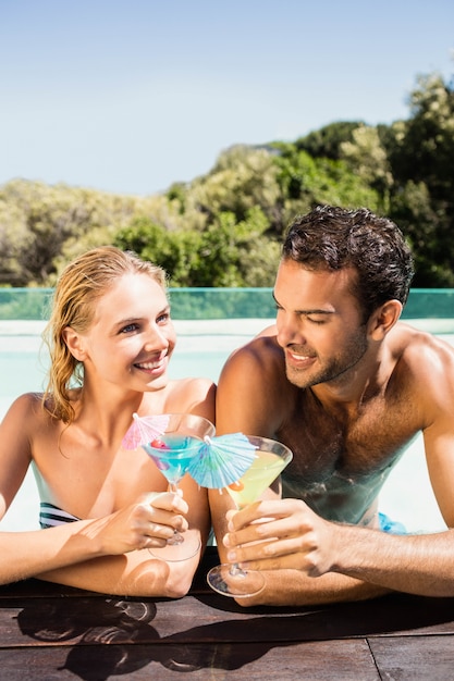 Casal feliz, apoiando-se na borda da piscina e brindar com cocktails em um dia ensolarado