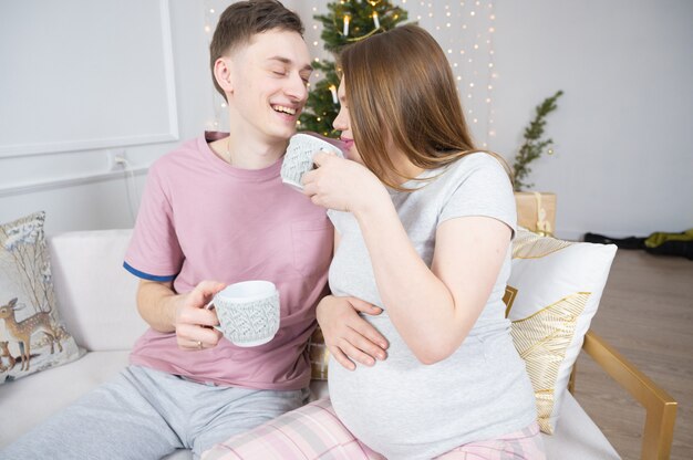 Casal feliz bebendo chá ou café em casa à noite, sobre as luzes da árvore de Natal no fundo. Conceito de férias, lazer e pessoas.