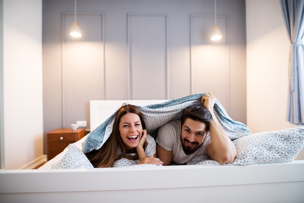 Casal feliz brincalhão olhando para a câmera enquanto estava deitado na cama, debaixo de um cobertor.