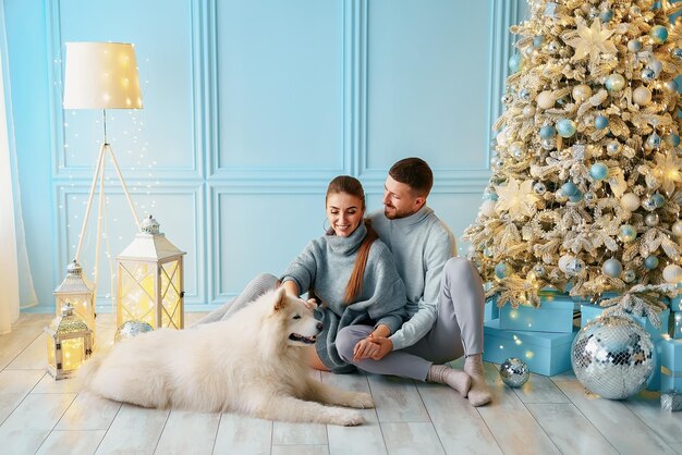 Casal feliz com um cachorro sentado perto da árvore de Natal em casa, roupas e suéteres domésticos