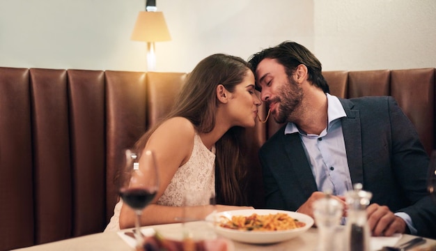 Casal feliz compartilhando comida e beijo para jantar abraço ou romance de relacionamento à noite no restaurante Homem e mulher inclinam-se com a boca de macarrão para beijos íntimos em um jantar requintado no dia dos namorados