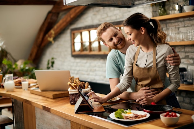 Casal feliz conversando enquanto usa o touchpad e prepara comida na cozinha