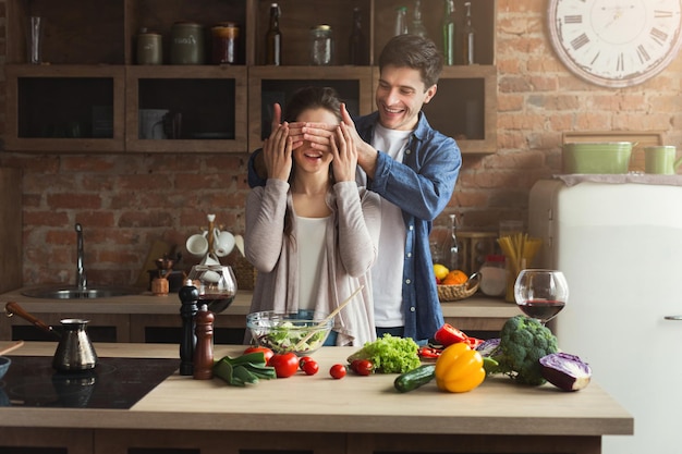 Casal feliz cozinhando alimentos saudáveis e se divertindo juntos na cozinha do sótão em casa. Mulher e homem brincando de esconde-esconde. Preparando salada de legumes.